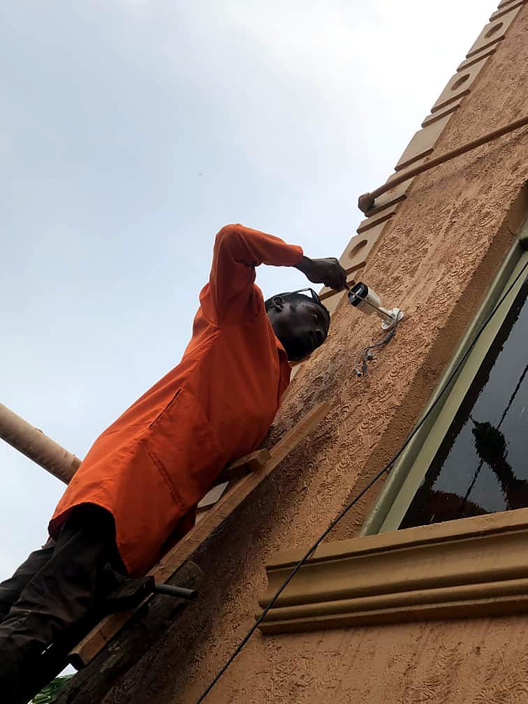 Technician in orange overalls mounting a CCTV bullet camera high on a building facade
